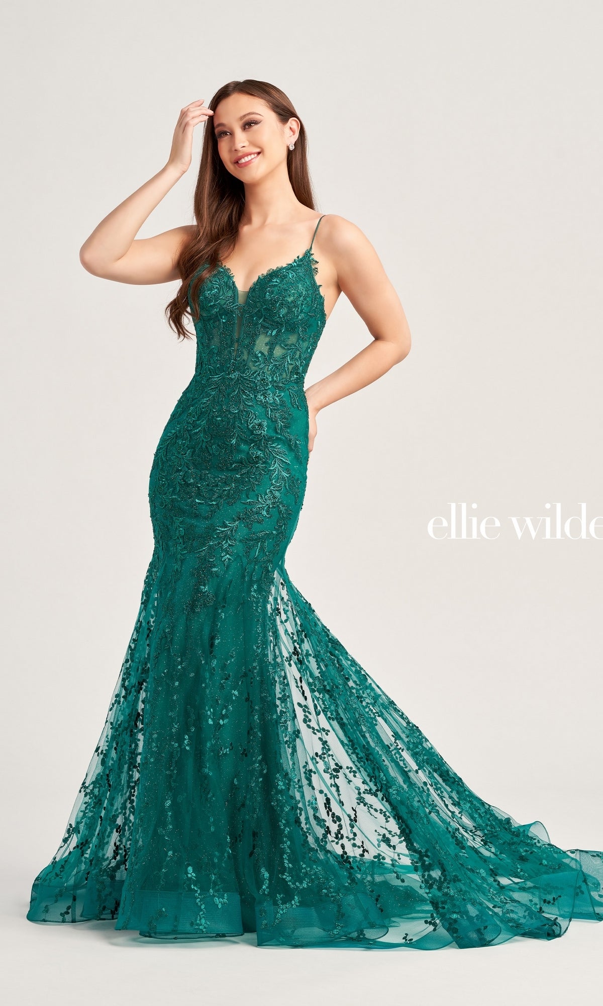 A woman poses against a plain background wearing the Ellie Wilde Beaded-Tulle Long Prom Dress EW35010—an emerald green, fitted gown with thin straps, beaded embroidery, a flared train, and intricate detailing.